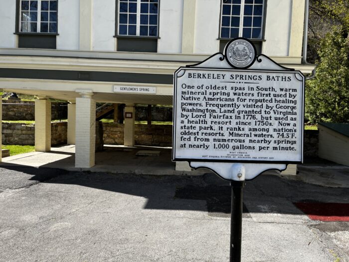 An official sign referencing the history of Berkeley Springs Baths stands in a park.