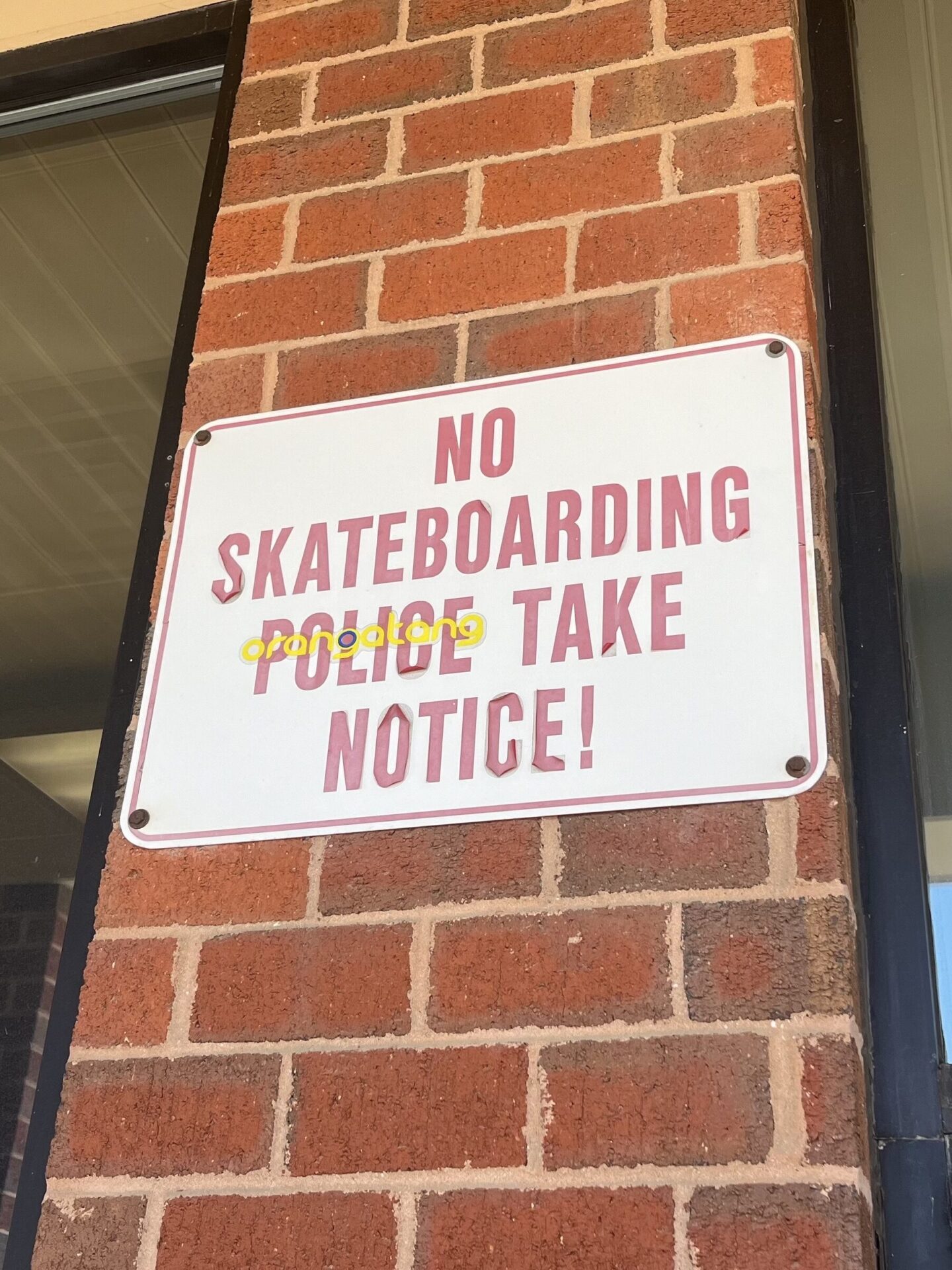 A red and white sign sits on the red-brick exterior of a building with tall glass pane windows. The sign reads, "NO Skateboarding. Police Take Notice!" A yellow sticker has been placed atop the word "police."