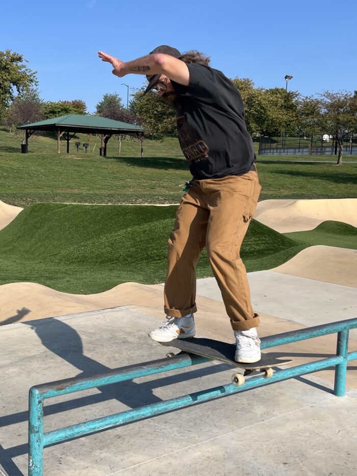 A man in brown pants and a black shirt rides a skateboard atop a low-to-the-ground rail. Skateboard ramps and park slides are visible in the background.