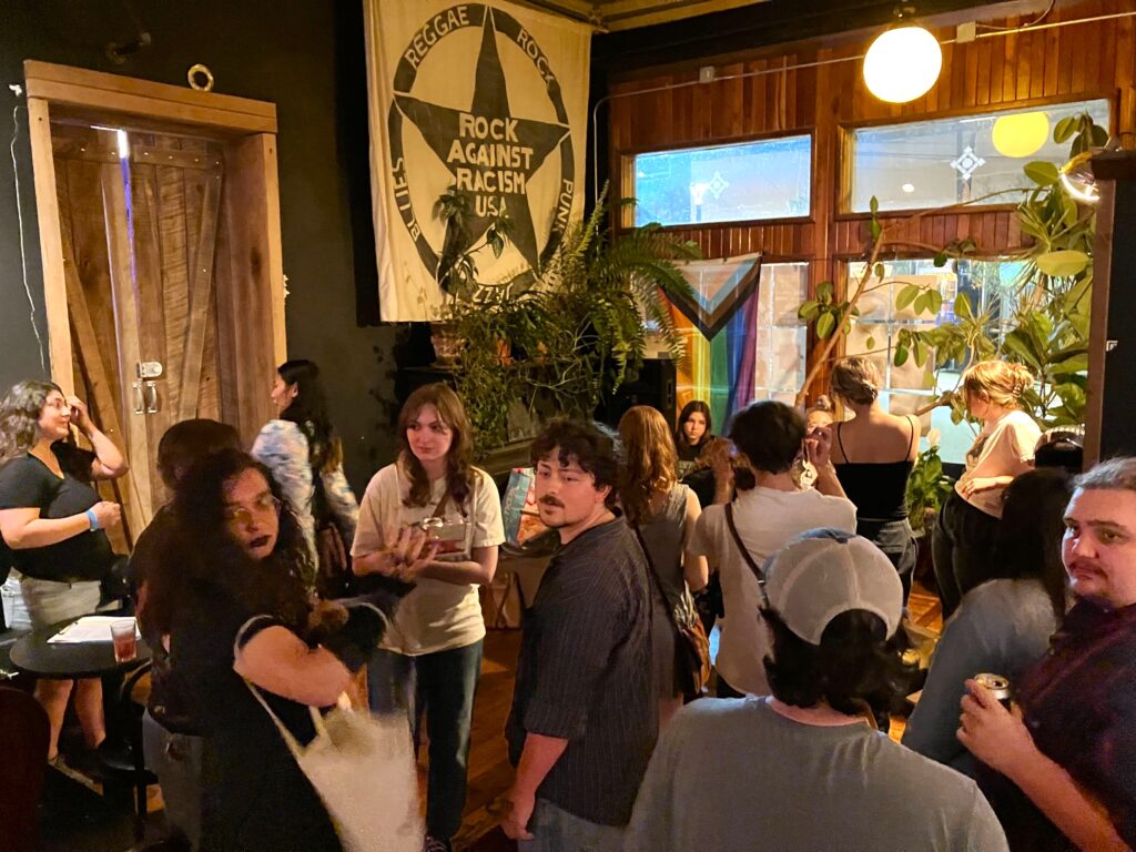 A crowd of people stand in the corner of a room, one wall of which is a wall of windows. On the far wall is a banner with a large star printed on it, with the words "Rock Against Racism USA". An LGBTQIA flag is hanging in one of the windows.