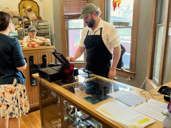 A man wears a black apron over a white collared shirt while reaching towards a touchpad on a glass-topped counter. In front of him stands a woman wearing a white dress with a floral pattern and a teal sweater with her hair in a messy bun. In the background another woman wearing a ball cap stands at a higher counter. On the glass-top counter can be seen a form with a blank table with yellow highlights.