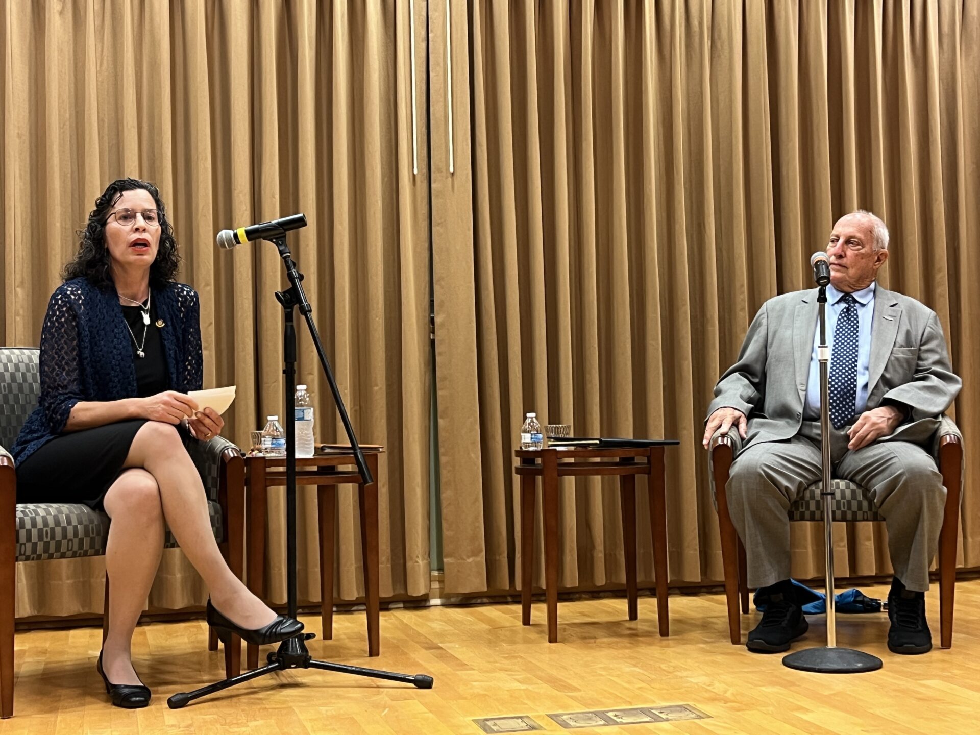 A woman and a man sit on a stage, facing an audience off screen. They speak into microphones. The women is speaking to a member of the audience.