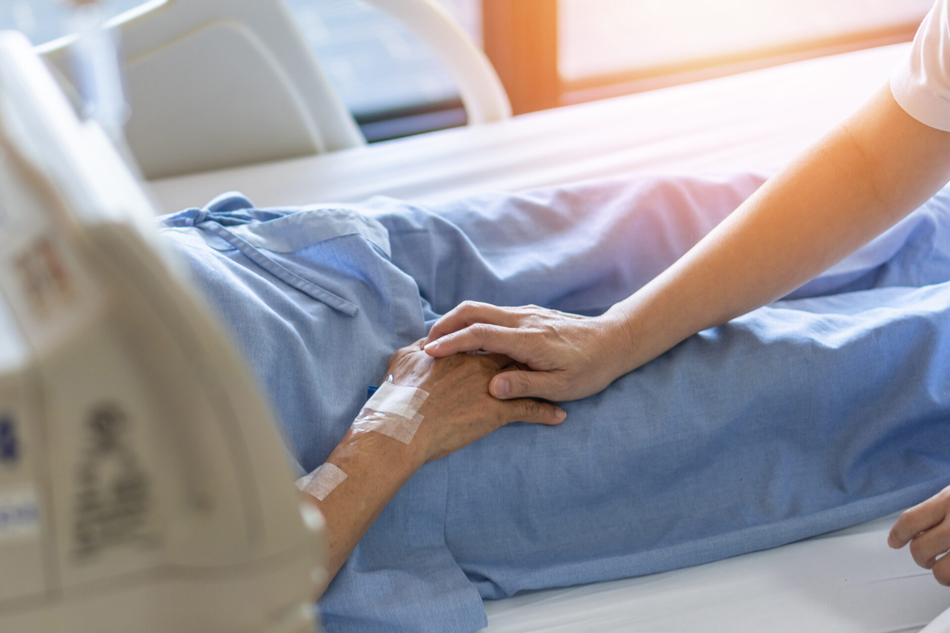 A patient lays in a hospital bed wearing blue scrubs. Sunlight streams in the room as a hand with a white sleeve reaches to hold the hand of the patient.