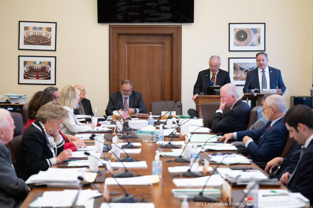 shot long table with lawmakers on all sides and two men speaking at a podium on the far end