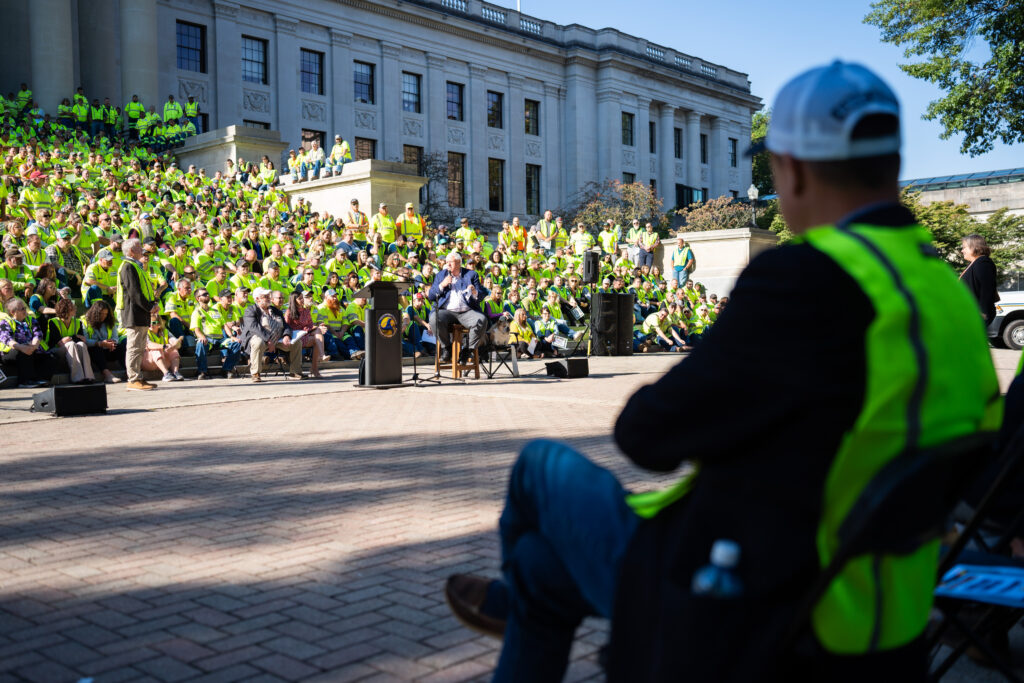 A man wearing a bright green construction vest is seen in the foreground of the image. He is seated with one leg crossed over the other and arms crossed over his chest as he listens to a man wearing a suit and standing in front of the West Virginia State Capitol speak. Surrounding the man in a suit are people wearing the same construction vests.