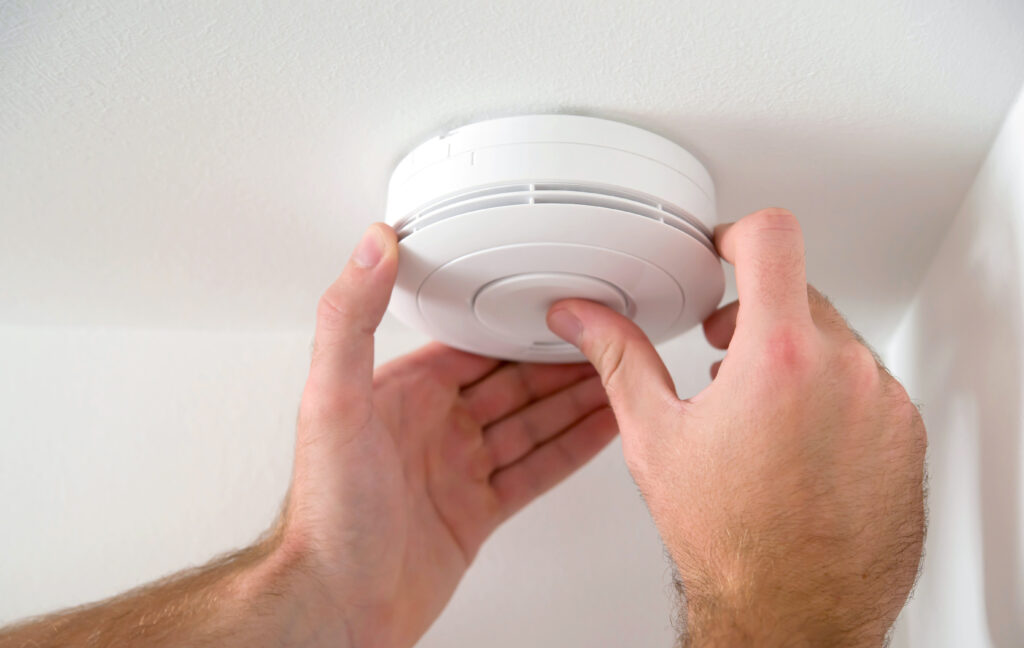 A pair of Caucasian hands reach and grab a smoke detector on the ceiling. The walls and ceiling are also white.