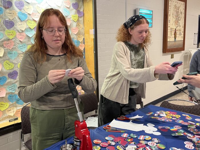 Two young women stand behind a table with a blue tablecloth. The woman on the left stands behind a red metal button press, and holds up a newly made button, looking down at it. The woman on the right is using her phone to scan a QR code being extended by an event attendee.