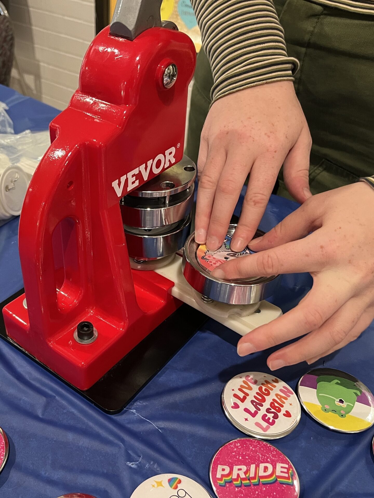 A red metal button press sits atop a table with a blue tablecloth. Colorful buttons sit beside it. A person's hand is pulling a newly finished button out of the press.