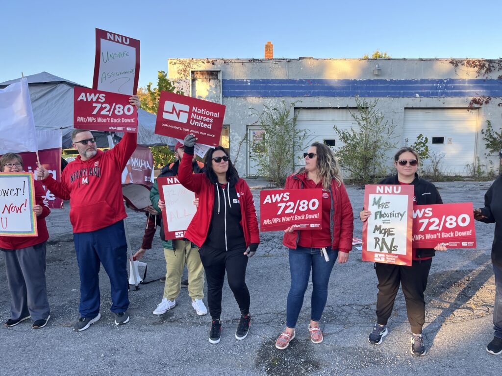 People in red shirt stand along a roadside, holding signs with different slogans in favor of flexible scheduling policies for nursing.