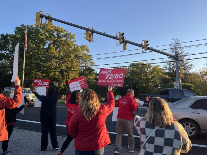 People wearing red stand along a road and hold signs up to passing cars. The signs read various slogans in favor of flexible scheduling policies for nurses.