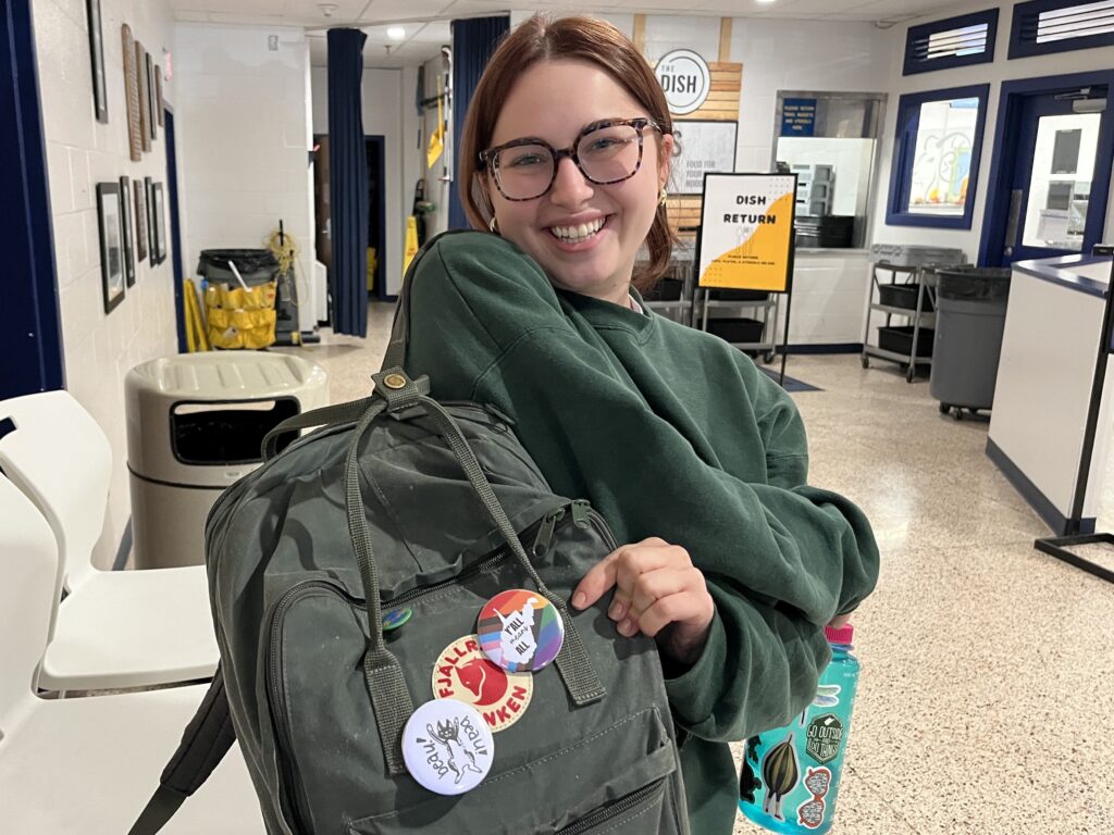 A young woman in a green sweatshirt stands in a college dining hall. She is holding a green backpack and gesturing toward a rainbow pin on it.