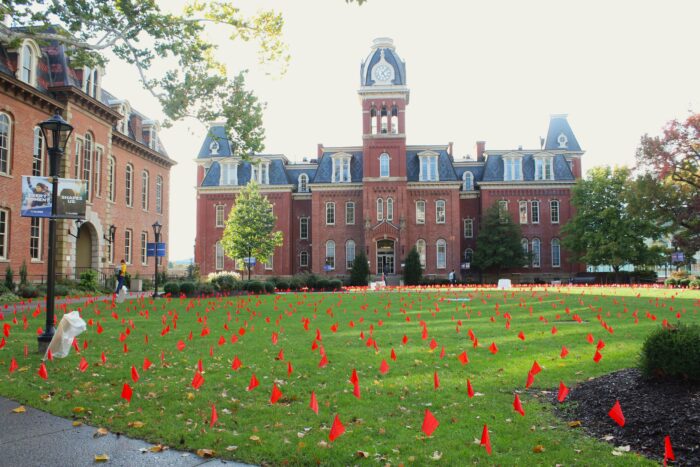 Red flags cover the green lawn in front of a brick building. The building has a large clock tower at its center that dominates the frame.