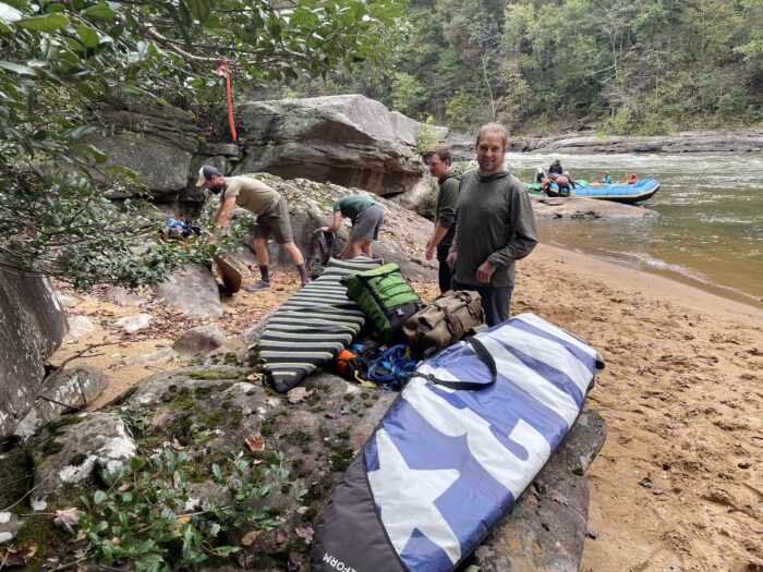 a group of men stand around a sandy river beach with surfboards in bags.