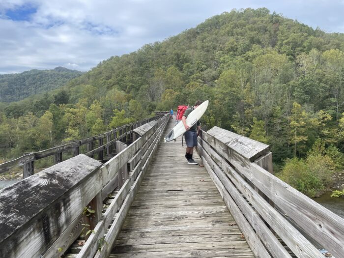 A man holds a surfboard on a small wooden bridge.