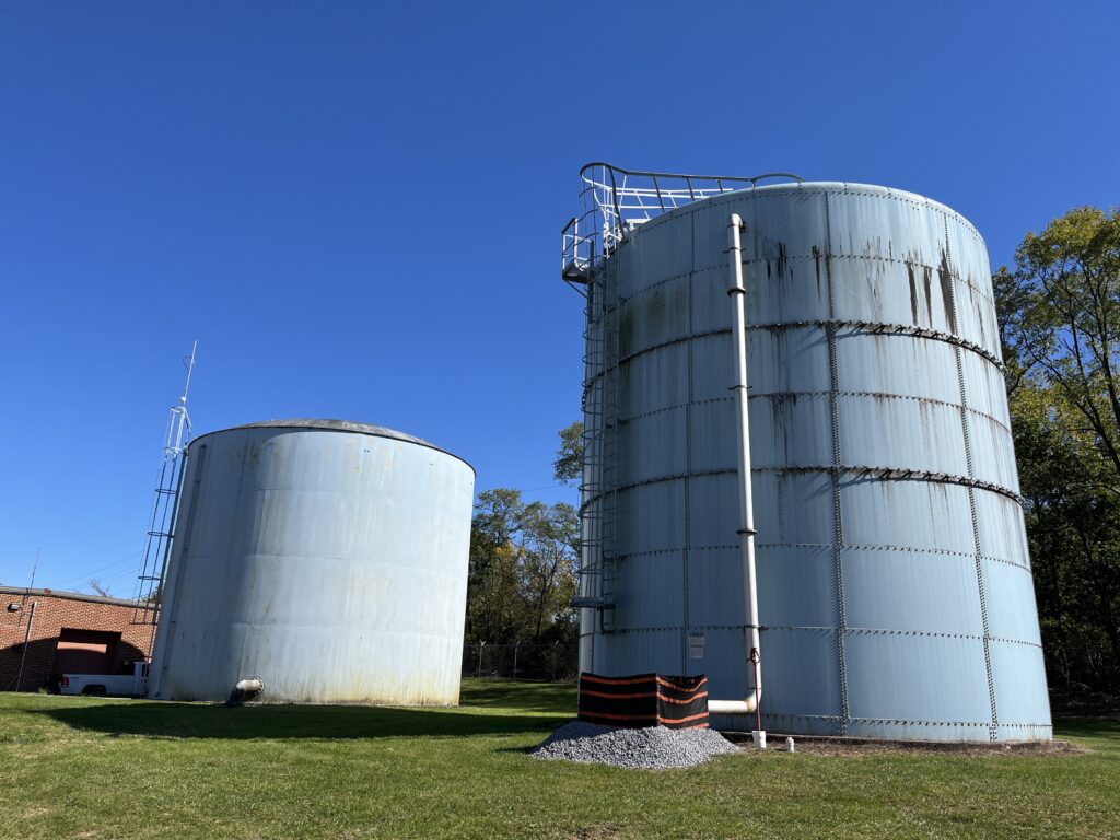 Two tall light blue water tanks with brown staining on their sides stand tall in the grass. Pipes protrude out from them, and a brick building sits in the background. The sky is blue overhead.