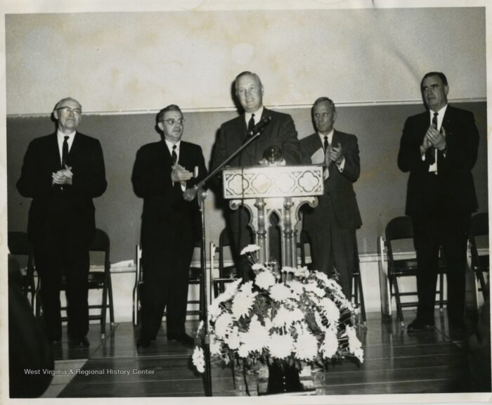 A black and white image of a man standing at a podium, flanked by two men on either side