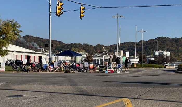 People holding signs picket along the roadside at the entrance to a facility.