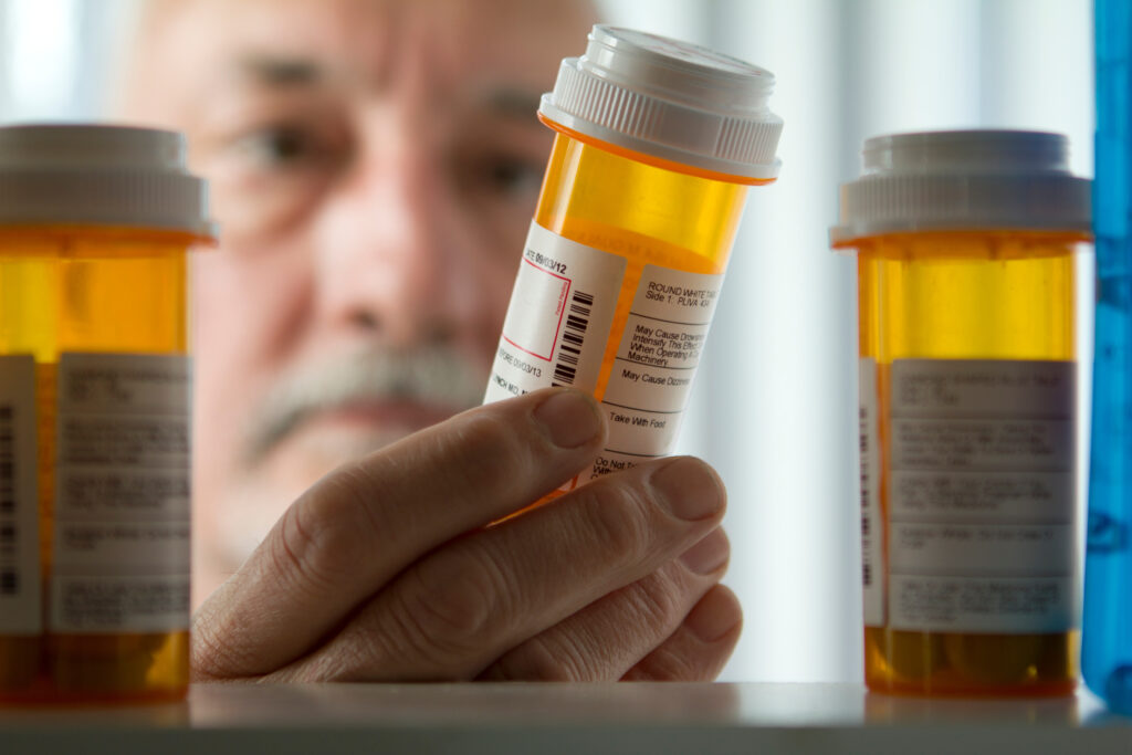 An older man with a grey and white mustache reaches into a medicine cabinet to read the label on a prescription bottle.