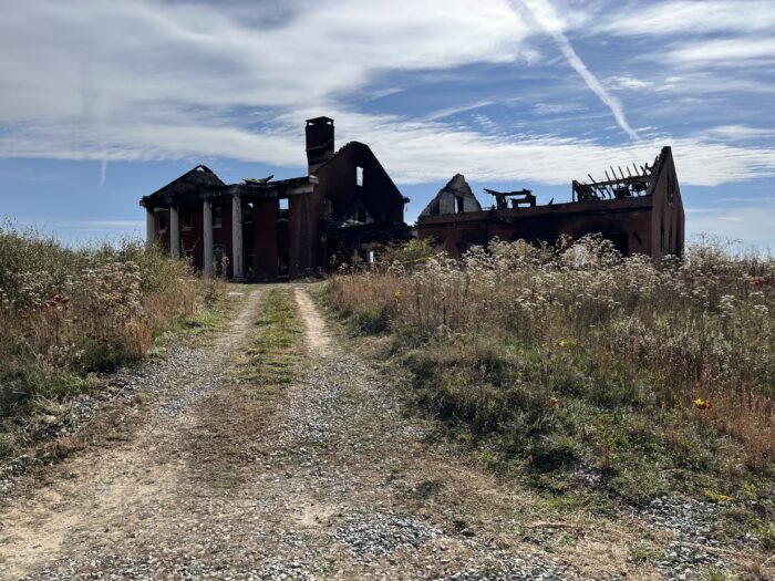 A large burnt-out structure is seen on a gravel and dirt road in front of a blue sky with clouds.