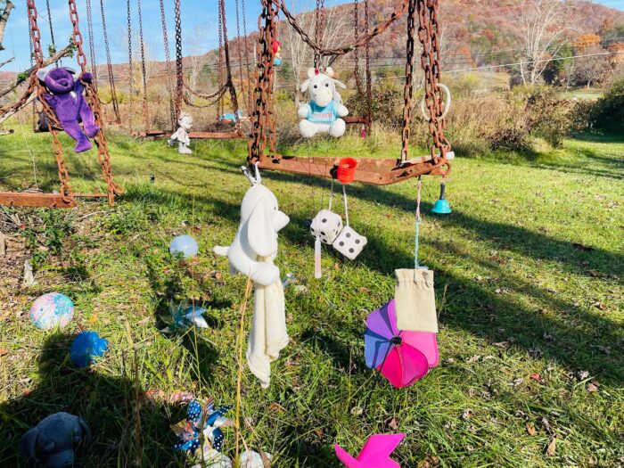 a rusty metal swing sits still with stuff animals tied to it. On the ground there are windchimes and other stuffed animals.