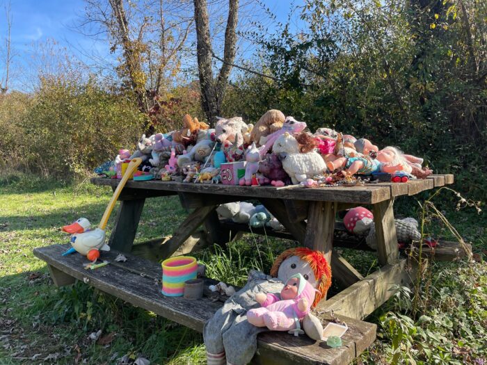 a outside picnic table full of stuffed animals that are dirty from the elements. Its a sunny day and the stuffed animals create a vibrant display.