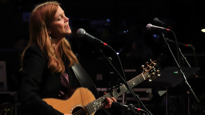 A woman with auburn hair sings into a mic and plays the guitar.