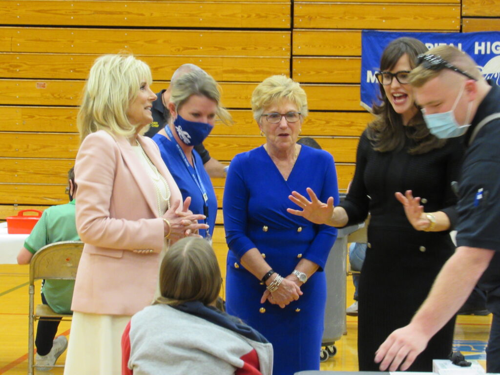 A woman in a blue dress stands a the center of a group in a wood-clad high school gymnasium.