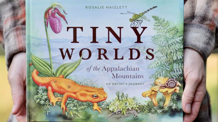 A woman holds a picture book with plants, insects and a salamander on the cover.