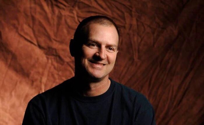 A man in charcoal t-shirt smiles in front of a brown backdrop.