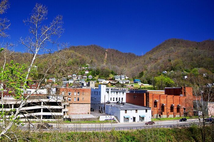 A blue sky hangs over two mountain peaks. In the valley down below, buildings line city streets, viewed from an outcropping across a highway along the side of town.