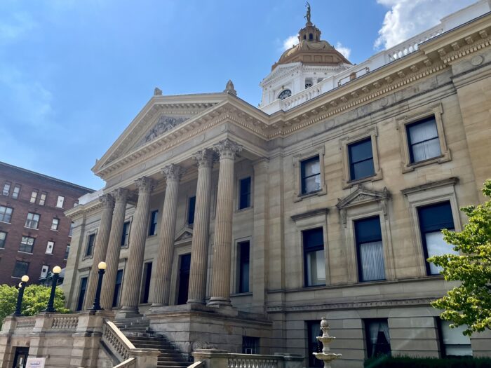 A tall building with pillars and long windows has a spire on top, sitting against a clear blue sky.