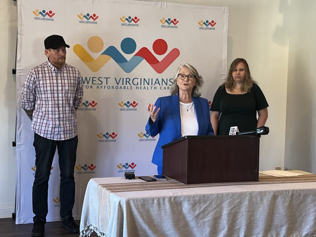 Three people stand behind a table and wooden speaking podium. A woman wearing a blue suit is speaking while a man wearing a checkered shirt and hat stands to the left and a woman with blonde hair wearing a green dress stands to the right.