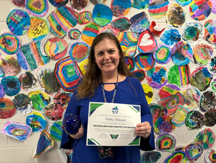 A woman with long brown hair is seen standing in front of a colorful wall featuring children's art. The woman is wearing a blue dressy shirt, holding a certificate, and a blue glass apple. She is smiling.