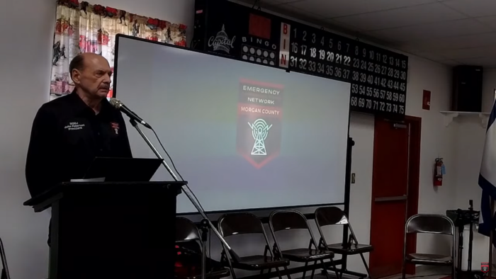 A man speaking into a microphone stands in front of a screen that displays a logo reading "Emergency Network Morgan County."