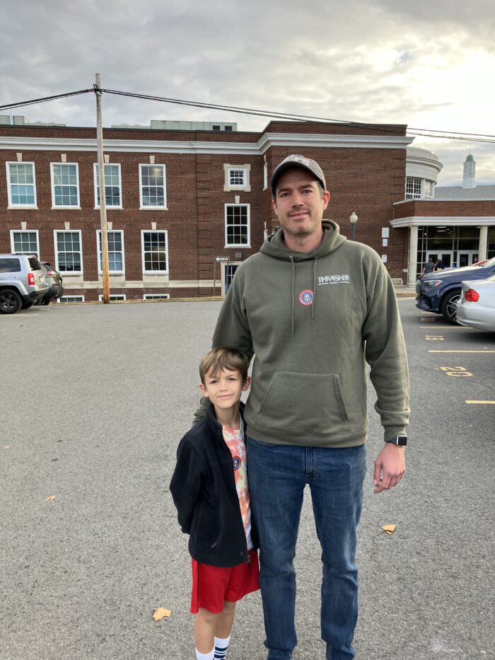 A man wearing an olive green hoodie over blue jeans and a baseball cap stands next to a boy wearing red shorts and a dark blue zip sweater over a tie dye shirt stand in the parking lot in front of a brick building with many double hung windows in its facade. An "I voted" sticker is displayed in the center of the man's chest