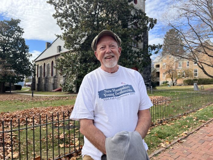 A man in a hat and a white shirt with writing on it "Young Democrats of New Hampshire" standing on a red brick driveway and smiling at the camera. Behind it behind a short black fence sits a stone church and courtyard.