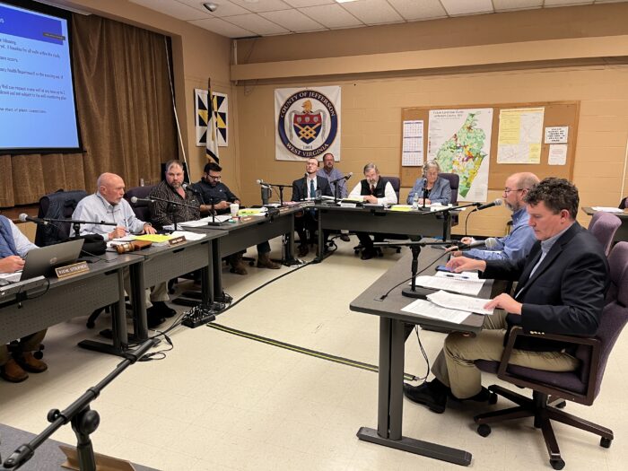 Two men sit at a desk in front of rows of audience members. They are speaking into microphones and facing a desk filled with commissioners identified by name placards.