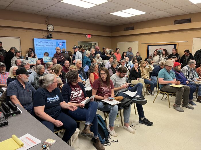 A crowd of people sit in rows of chairs facing forward, some holding hand-written notes and other documents and others holding hand-painted signs. The room is filled beyond capacity, and several people are watching in through the doorway.