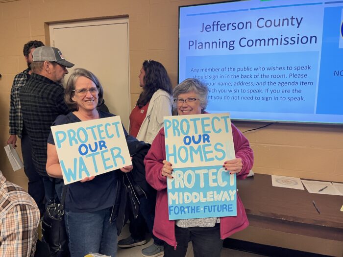 Two women stand in front of a circle of people chatting and a television screen that reads "Jefferson County Planning Commission." They are smiling at the camera and holding up hand-painted blue and white signs that read "Protect Our Water" and "Protect Our Homes."