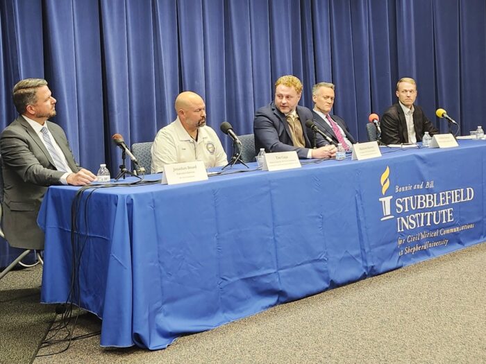 A panel of men in suits sit at a table with a blue tablecloth and speak into microphones.
