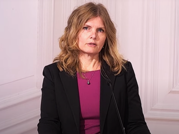 A woman in a magenta shirt and a blazer speaks into a microphone and looks into the camera.