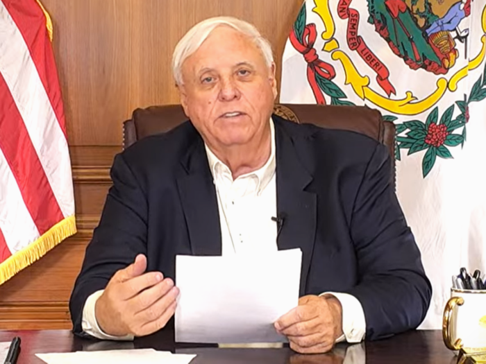 A man in a suit with an open collar sits at a desk and holds a piece of paper, gesturing with his empty hand. Behind him, the American and West Virginia flags are displayed.