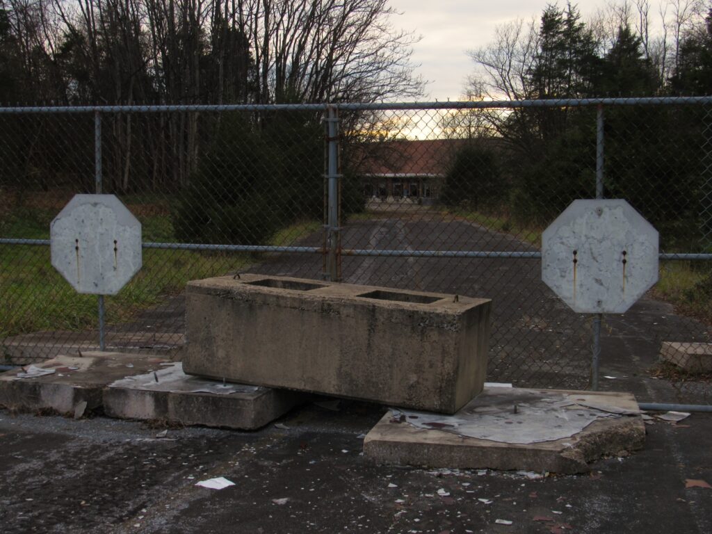 Cinderblocks and a chain-link fence block a paved path toward a worn-down building in the distance. Trees line the sides of the street.