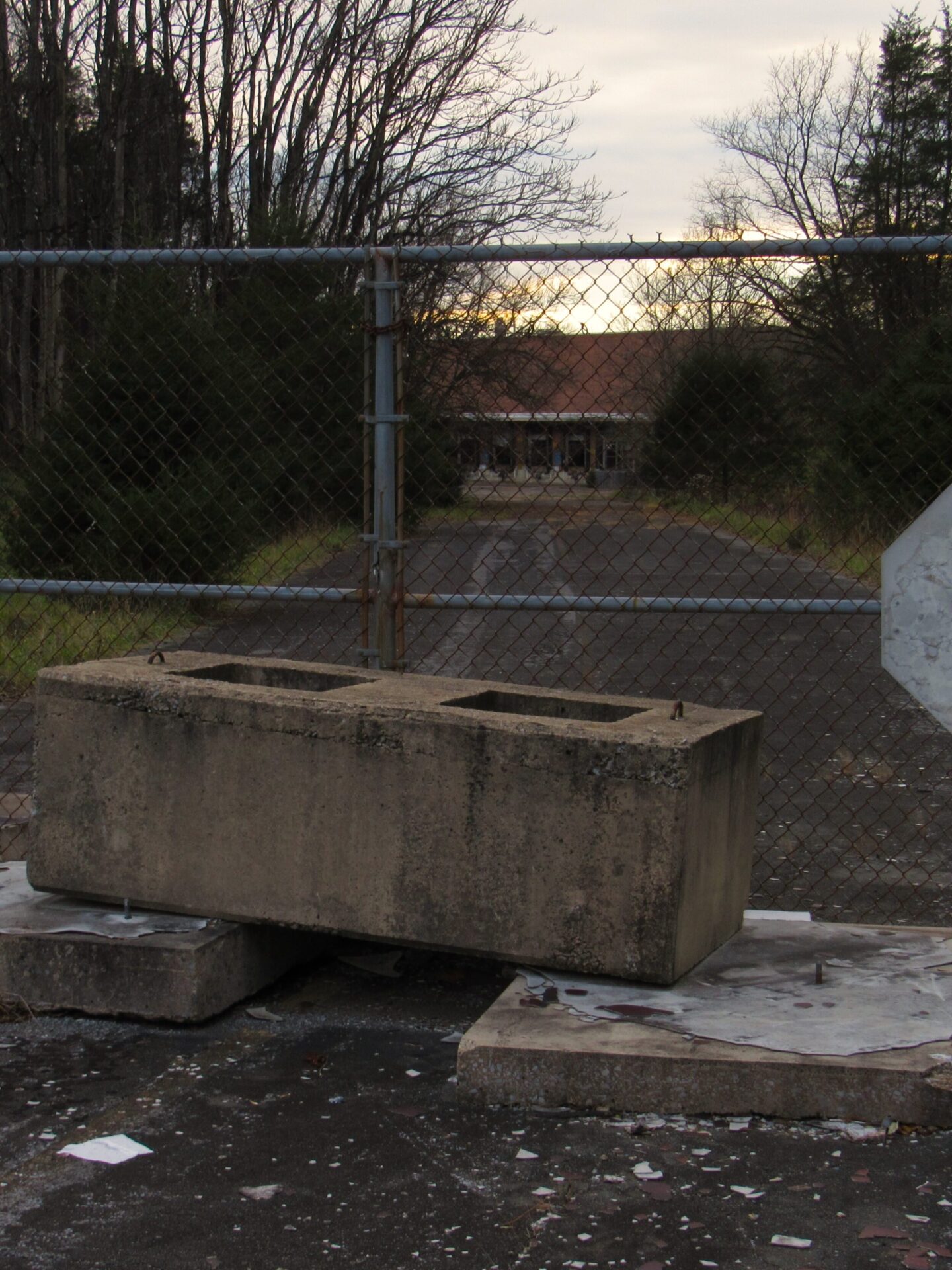 Cinderblocks and a chain-link fence block a paved path toward a worn-down building in the distance. Trees line the sides of the street.