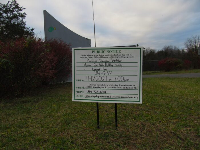 A sign in a lawn advertises a public comment period for plans on a proposed water bottling site in Middleway. It is placed in a lawn with trees in the background.