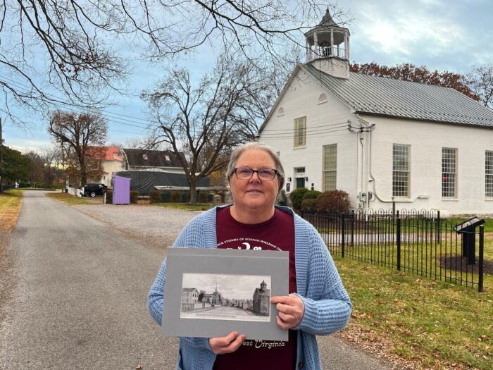 A woman stands on the edge of a street where vehicles are passing by, holding an old black-and-white drawing of the same street forward to the camera. She is looking toward the camera.