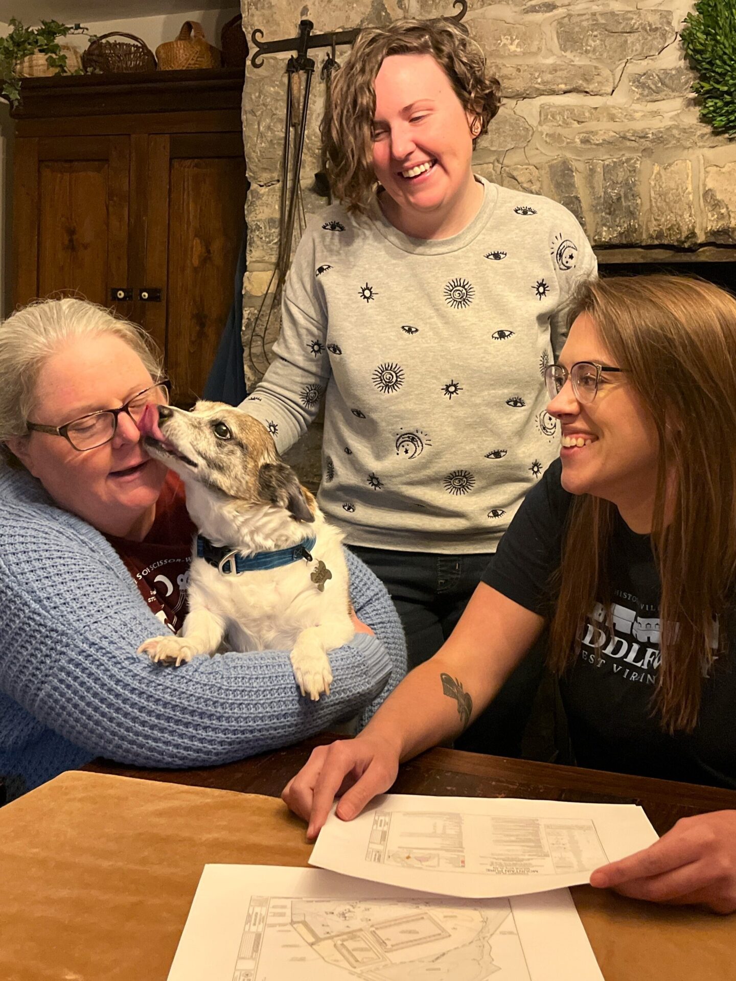 Three women sit at a wooden table and review printed out maps. The woman on the left holds a dog, who is licking her face. The other women look on and smile.