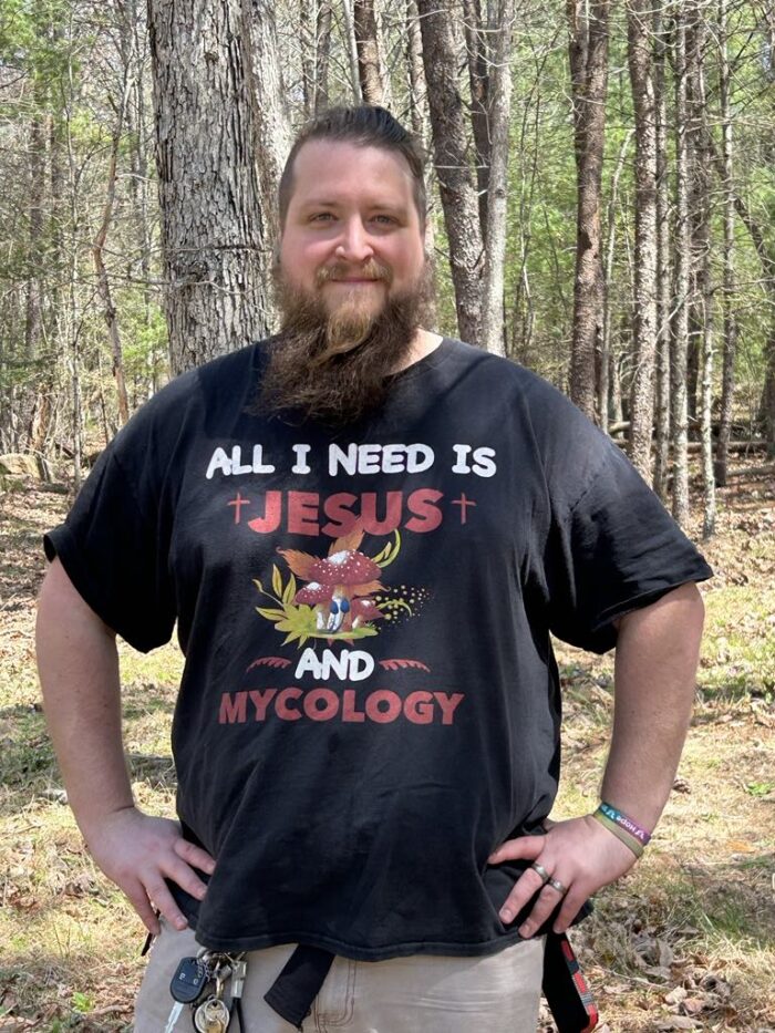 A man with brown hair and beard poses for a photo. Behind him are tall trees. He smiles and wears a black t-shirt that reads, "All I need is Jesus and mycology."