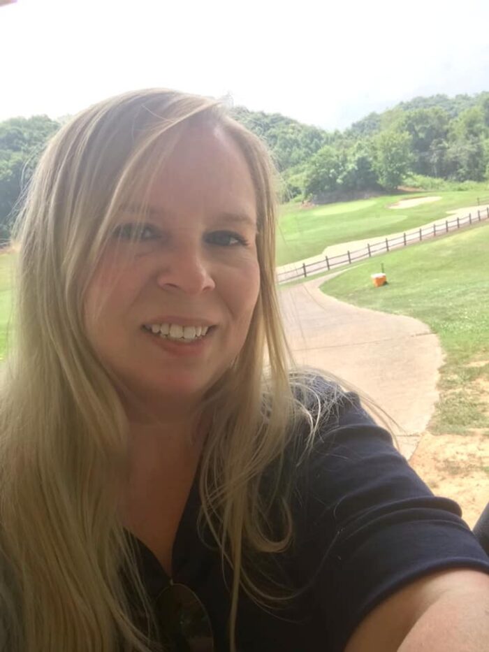 An adult woman smiles for the camera as she takes a selfie. Behind her are green trees and a driveway.