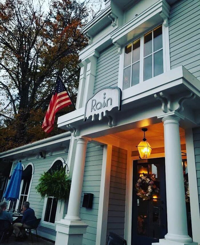 The outside of a building at dusk. Above the door is a sign that reads "Rain." There is an American flag hung on the porch and a wreath on the front of the door. The building is white with light blue panels.